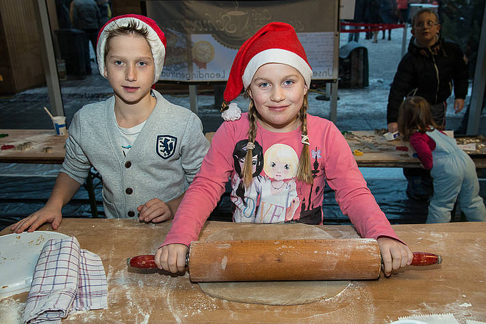 Zwei Kinder mit festlichen Weihnachtsmannmützen stehen an einem Tisch, wobei ein Mädchen mit einem Nudelholz Teig ausrollt, während der Junge zusieht.