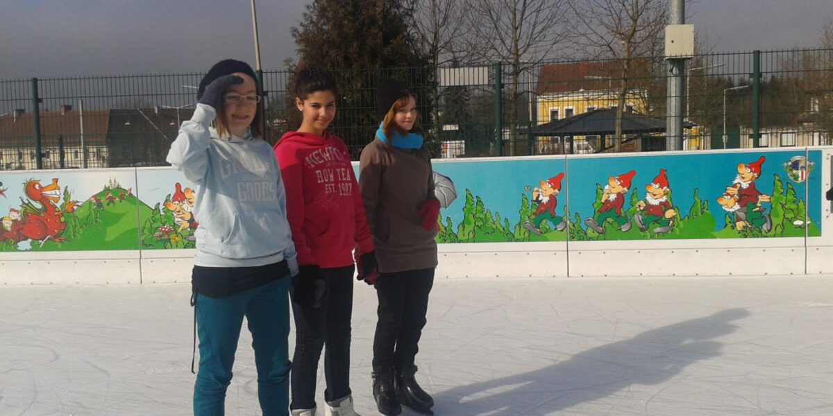 Drei Personen in Winterkleidung stehen auf einer Eislaufbahn im Freien mit einem bunten Gnom-Wandbild auf der Absperrung hinter ihnen.
