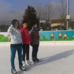 Drei Personen in Winterkleidung stehen auf einer Eislaufbahn im Freien mit einem bunten Gnom-Wandbild auf der Absperrung hinter ihnen.