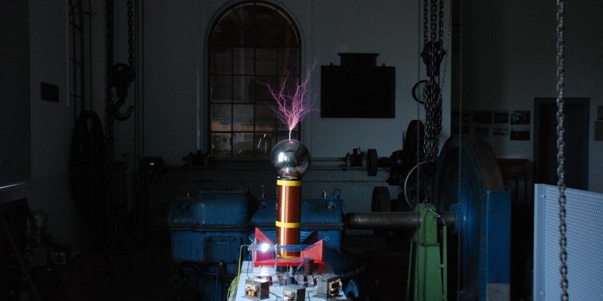 Eine Teslaspule, die violette elektrische Entladungen in einem schwach beleuchteten Industrieraum mit Maschinen und einem großen Fenster im Hintergrund abgibt.