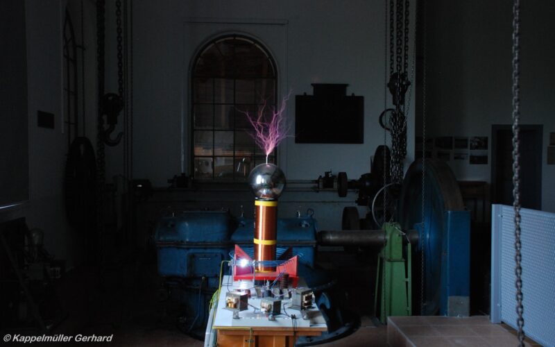 Eine Teslaspule, die violette elektrische Entladungen in einem schwach beleuchteten Industrieraum mit Maschinen und einem großen Fenster im Hintergrund abgibt.