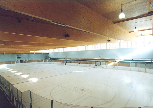 Eine leere Eishockeyhalle mit hölzernen Deckenbalken, durchsichtigen Anzeigetafeln und markierten Linien auf der Eisfläche.