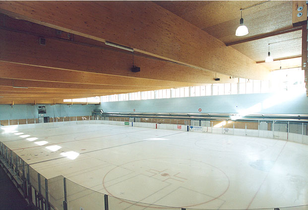 Eine leere Eishockeyhalle mit hölzernen Deckenbalken, durchsichtigen Anzeigetafeln und markierten Linien auf der Eisfläche.
