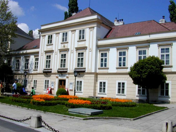 Ein zweistöckiges, weiß-beiges Gebäude im klassizistischen Stil mit symmetrischen Fenstern, einem zentralen Eingang und einem angelegten Garten mit Orangenblüten davor.