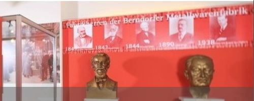 Eine Museumsausstellung mit Bronzebüsten vor einer roten Wand mit Porträts und Daten zu prominenten Persönlichkeiten der Berndorfer Metallwarenfabrik.