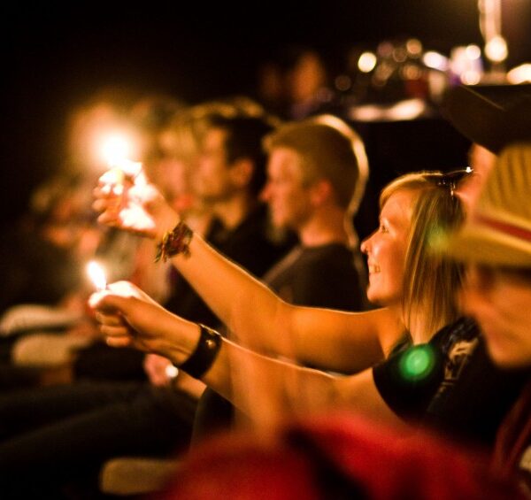 Eine Gruppe von Menschen, die in einem dunklen Raum sitzt, hält brennende Feuerzeuge in die Höhe und blickt nach vorne, einige lächeln und schauen aufmerksam zu.