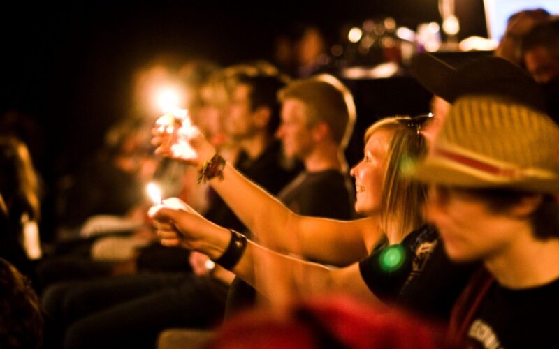 Eine Gruppe von Menschen, die in einem dunklen Raum sitzt, hält brennende Feuerzeuge in die Höhe und blickt nach vorne, einige lächeln und schauen aufmerksam zu.