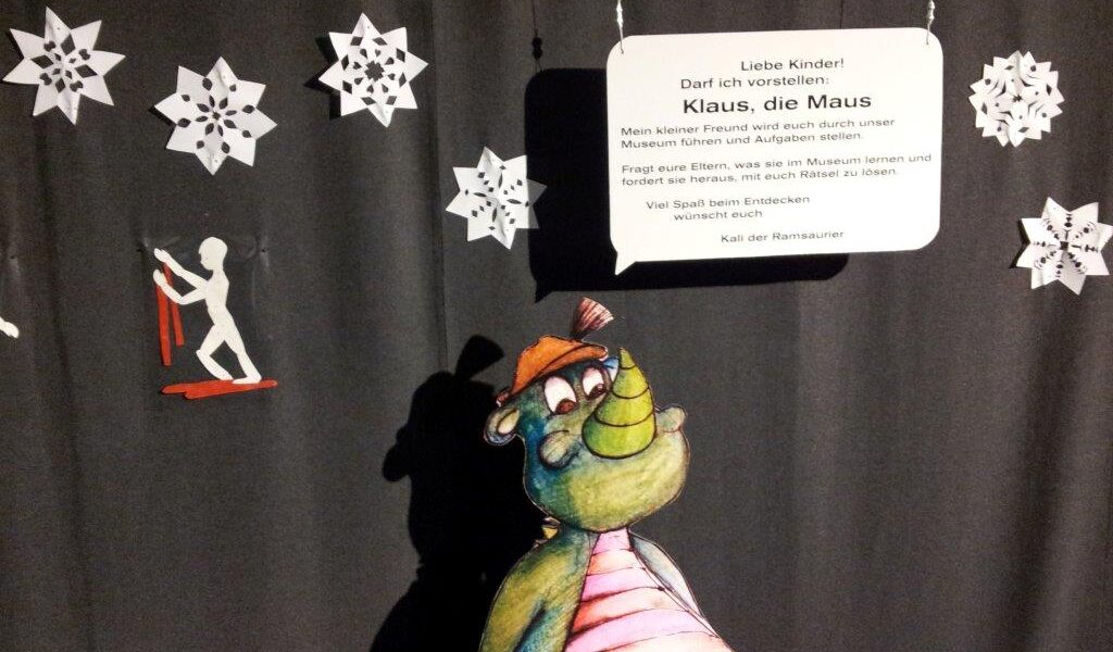 Ein Cartoon-Dinosaurier steht vor schwarzen Vorhängen, die mit weißen Papierschneeflocken und einem Schild mit der Aufschrift "Klaus, die Maus" verziert sind.