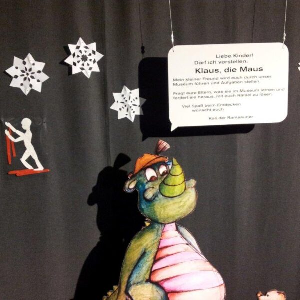 Ein Cartoon-Dinosaurier steht vor schwarzen Vorhängen, die mit weißen Papierschneeflocken und einem Schild mit der Aufschrift "Klaus, die Maus" verziert sind.