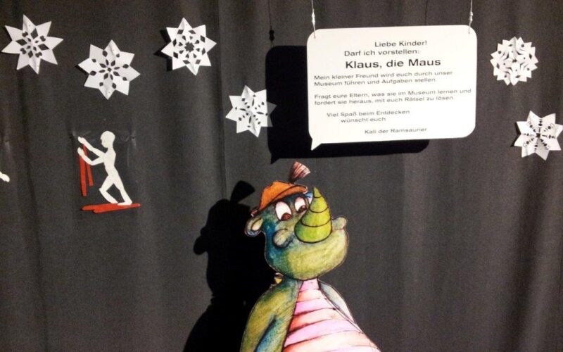 Ein Cartoon-Dinosaurier steht vor schwarzen Vorhängen, die mit weißen Papierschneeflocken und einem Schild mit der Aufschrift "Klaus, die Maus" verziert sind.