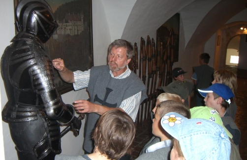 Ein Mann erklärt einer Gruppe von Kindern in einem Museum oder einer historischen Ausstellung eine mittelalterliche Rüstung.