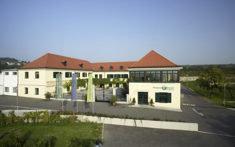 Ein moderner Gebäudekomplex mit roten Ziegeldächern, cremefarbenen Wänden und großen Fenstern, umgeben von einem Parkplatz und Grünflächen unter einem klaren Himmel.