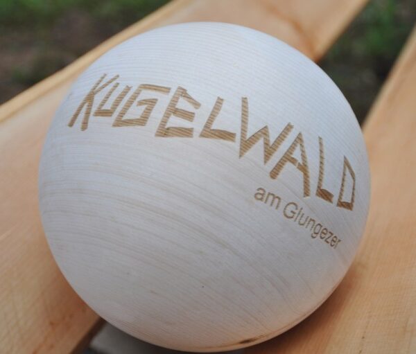 Eine glatte Holzkugel mit dem eingravierten Schriftzug "KUGELWALD am Glungezer", die auf einer hellen Holzfläche ruht.