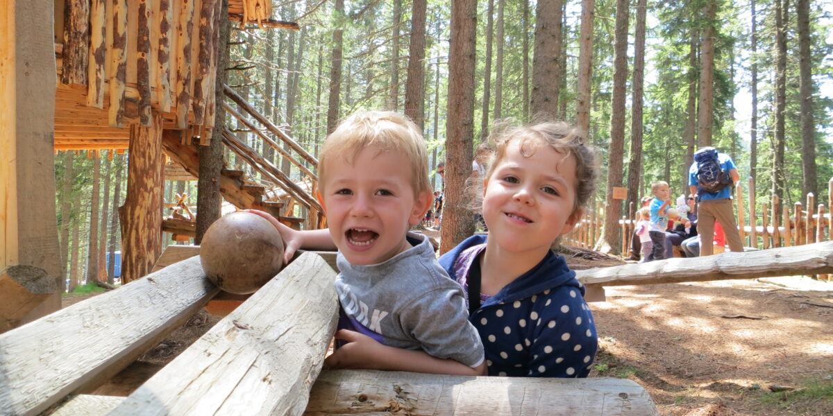 Zwei lächelnde Kinder lehnen auf Holzbalken vor einer rustikalen Hütte im Wald, im Hintergrund sind zwischen hohen Bäumen weitere Menschen zu sehen.