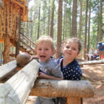 Zwei lächelnde Kinder lehnen auf Holzbalken vor einer rustikalen Hütte im Wald, im Hintergrund sind zwischen hohen Bäumen weitere Menschen zu sehen.
