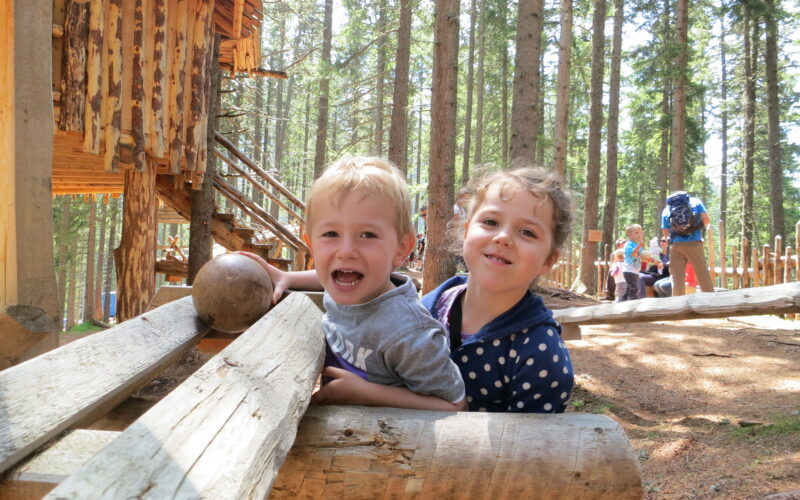 Zwei lächelnde Kinder lehnen auf Holzbalken vor einer rustikalen Hütte im Wald, im Hintergrund sind zwischen hohen Bäumen weitere Menschen zu sehen.
