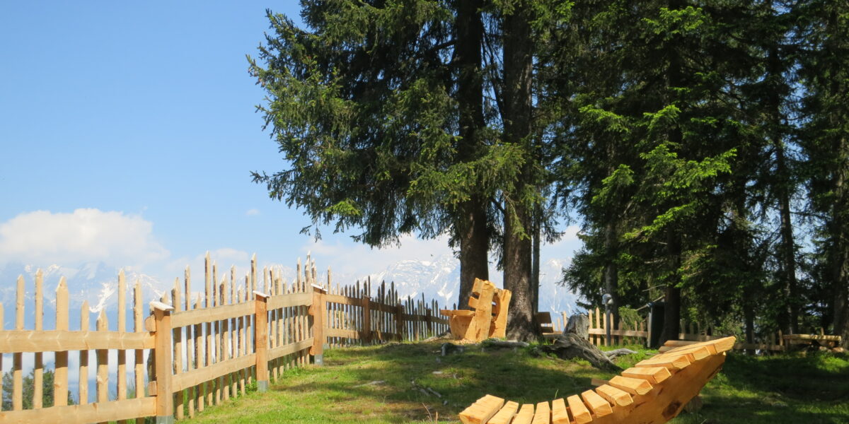 Ein hölzerner Liegestuhl steht auf grünem Gras in der Nähe eines rustikalen Holzzauns und hoher immergrüner Bäume unter einem klaren blauen Himmel. In der Ferne sind schneebedeckte Berge zu sehen.