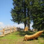 Ein hölzerner Liegestuhl steht auf grünem Gras in der Nähe eines rustikalen Holzzauns und hoher immergrüner Bäume unter einem klaren blauen Himmel. In der Ferne sind schneebedeckte Berge zu sehen.