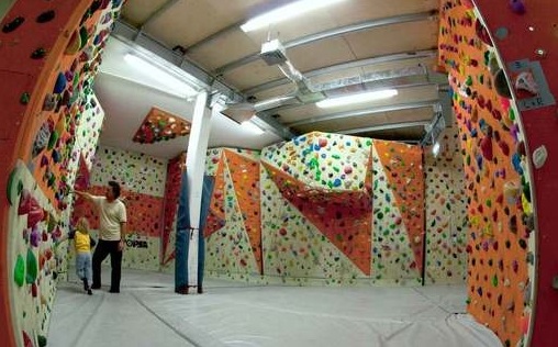 Eine Indoor-Kletterhalle mit gepolsterten Böden, bunten Klettergriffen und zwei Personen, die an einer Kletterwand stehen.