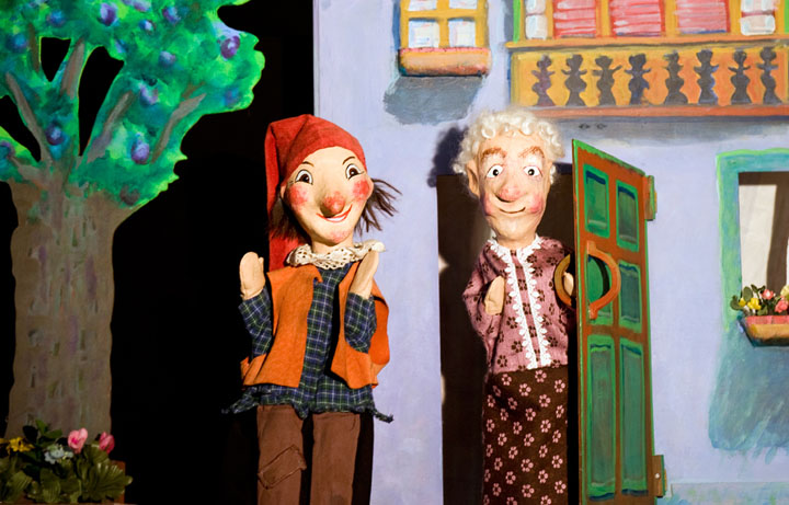 Zwei Puppen, die ein Mädchen und eine ältere Frau darstellen, stehen vor einem bemalten Haus mit einem Baum und Blumen im Hintergrund.