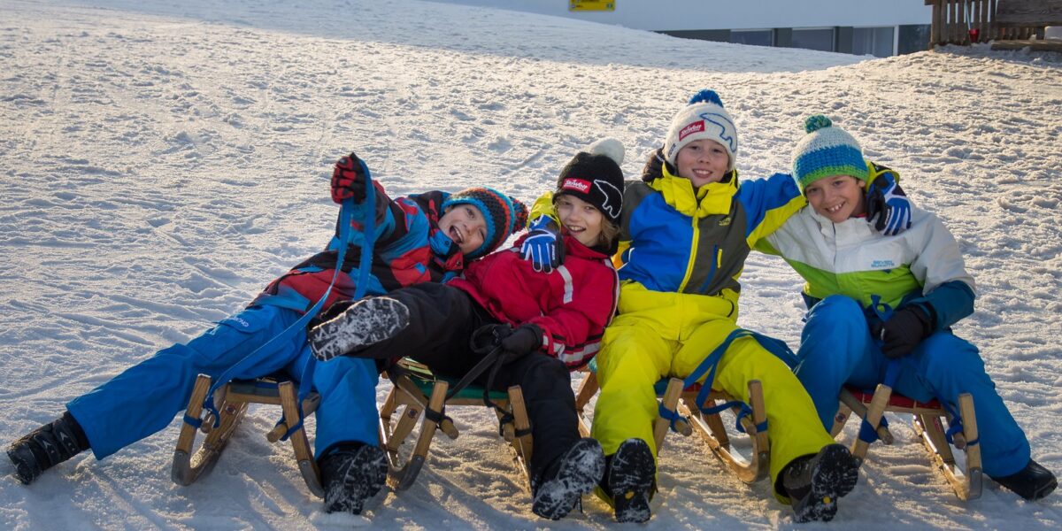 Vier Kinder in Winterkleidung sitzen auf Schlitten im Schnee, lächeln und posieren für das Foto mit ihren Armen umeinander.