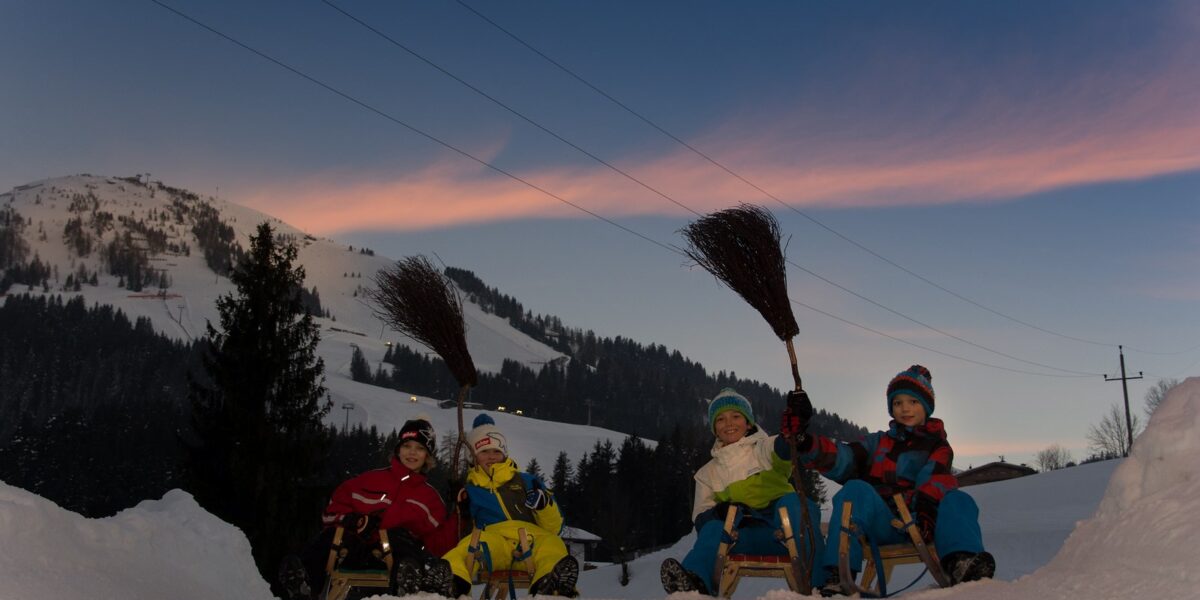 Vier winterlich gekleidete Kinder sitzen auf Schlitten im Schnee und halten Besen in die Höhe. Im Hintergrund sind verschneite Berge und Bäume bei Sonnenuntergang zu sehen.