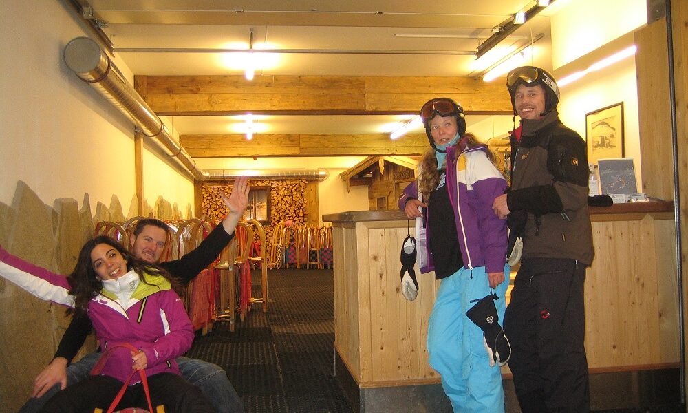 Vier Personen in Skikleidung posieren in einem Innenraum; zwei sitzen auf einem Schlitten, und zwei stehen mit Helmen und Brillen an einem Tresen. Hinter ihnen sind Schlitten an der Wand aufgereiht.