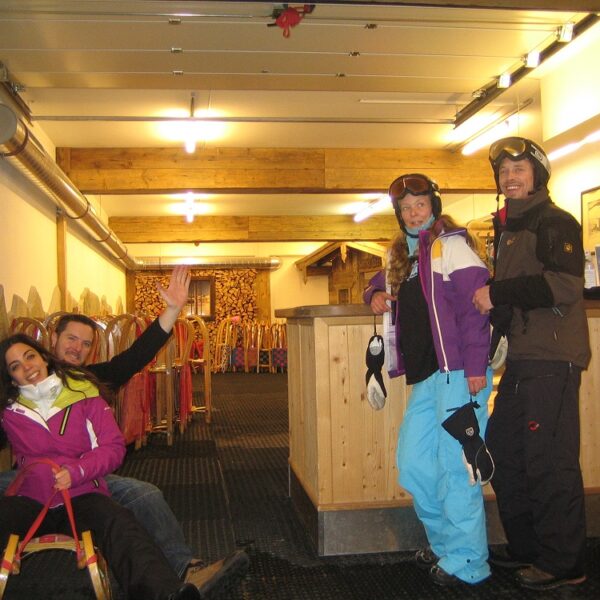 Vier Personen in Skikleidung posieren in einem Innenraum; zwei sitzen auf einem Schlitten, und zwei stehen mit Helmen und Brillen an einem Tresen. Hinter ihnen sind Schlitten an der Wand aufgereiht.