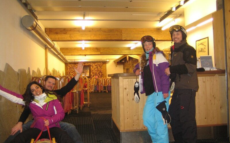 Vier Personen in Skikleidung posieren in einem Innenraum; zwei sitzen auf einem Schlitten, und zwei stehen mit Helmen und Brillen an einem Tresen. Hinter ihnen sind Schlitten an der Wand aufgereiht.