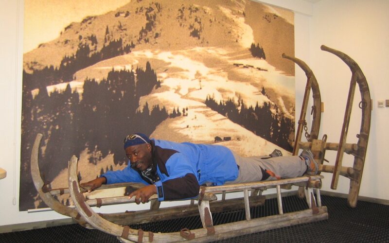 Ein Mann liegt mit dem Gesicht nach unten auf einem großen Holzschlitten im Haus und posiert, als würde er darauf fahren.