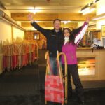 Zwei Personen stehen in einem Haus mit erhobenen Armen und lächelnd neben einem Holzschlitten. Im Hintergrund sind Reihen ähnlicher Schlitten aufgereiht. Die Umgebung scheint ein Verleih oder eine Ausrüstungsfirma zu sein.