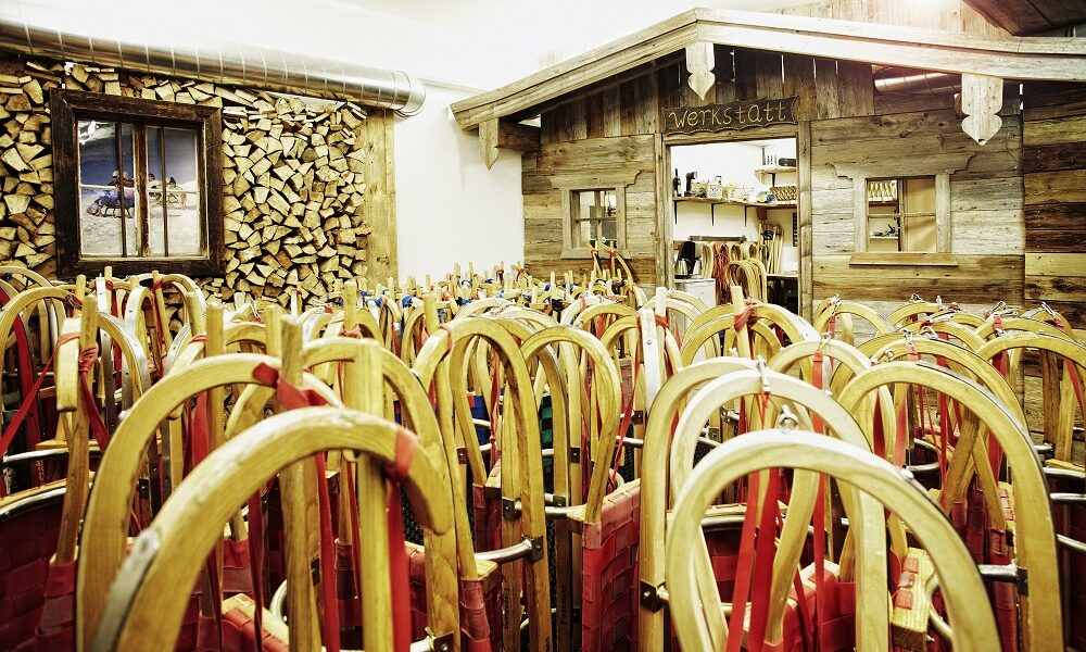 Ein Raum voller Holzschlitten mit roten Sitzen, aufrecht gestapelt neben einer rustikalen Holzwerkstatt und einer Wand mit gestapeltem Brennholz.