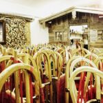 Ein Raum voller Holzschlitten mit roten Sitzen, aufrecht gestapelt neben einer rustikalen Holzwerkstatt und einer Wand mit gestapeltem Brennholz.