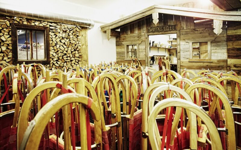 Ein Raum voller Holzschlitten mit roten Sitzen, aufrecht gestapelt neben einer rustikalen Holzwerkstatt und einer Wand mit gestapeltem Brennholz.