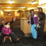 Vier Personen in Skikleidung posieren in einem Haus mit Schlitten; zwei sitzen auf einem Schlitten und zwei stehen neben einem Holztresen und zeigen die Daumen nach oben.