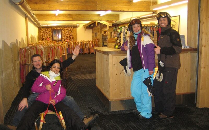 Vier Personen in Skikleidung posieren in einem Haus mit Schlitten; zwei sitzen auf einem Schlitten und zwei stehen neben einem Holztresen und zeigen die Daumen nach oben.