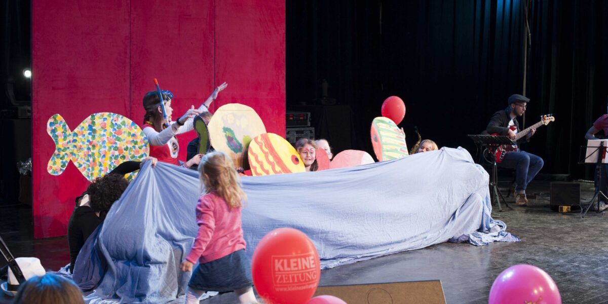 Kinder und Erwachsene treten auf der Bühne mit bunten Fischrequisiten und einem blauen Tuch auf, während ein Musiker nebenbei Gitarre spielt; Zuschauer mit Luftballons schauen zu.
