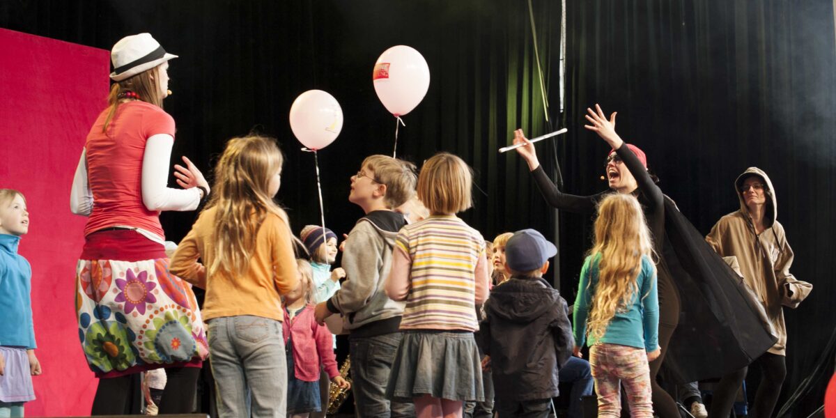 Eine Gruppe von Kindern versammelt sich auf einer Bühne und interagiert mit zwei Erwachsenen in Kostümen - einer in einem geblümten Rock, der andere in einem schwarzen Umhang -, während ein weiterer Erwachsener von der Seite zusieht.