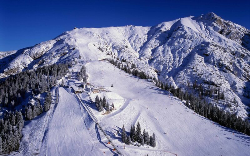 Schneebedeckte Skipisten auf einem Berg mit Skiliften, Bäumen und blauem Himmel im Hintergrund.