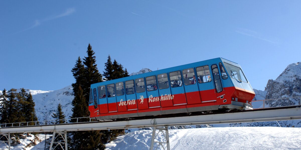 Eine rot-blaue Standseilbahn fährt auf einer Hochbahnstrecke durch eine verschneite Berglandschaft mit immergrünen Bäumen und strahlend blauem Himmel.