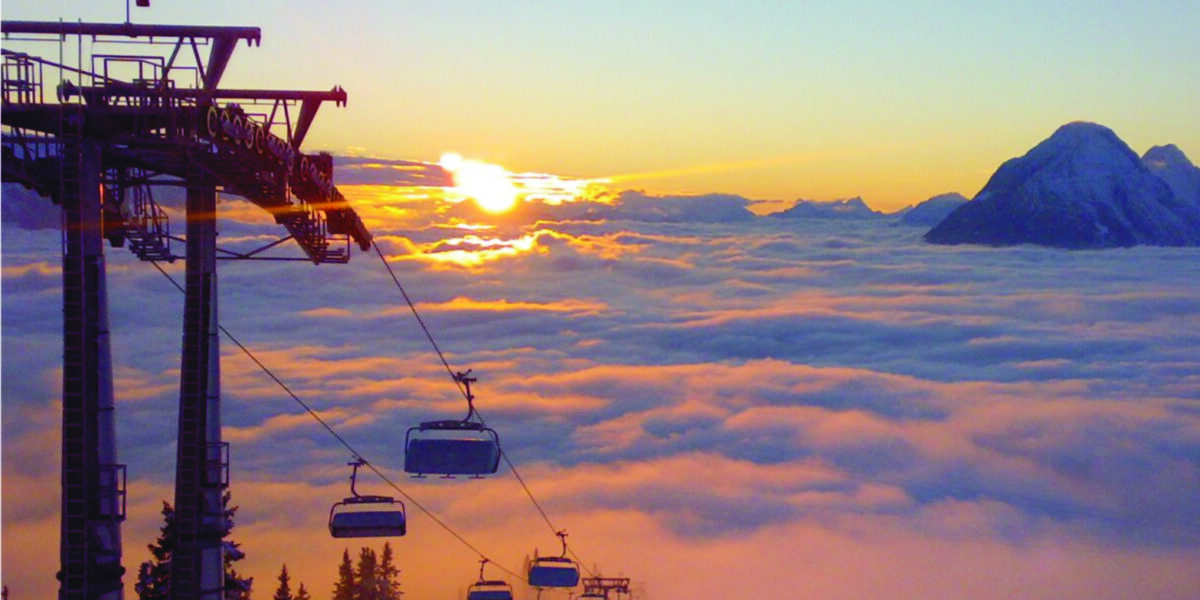 Über einem wolkenverhangenen Tal schwebende Skiliftsessel bei Sonnenaufgang, im Hintergrund sind die Berggipfel zu sehen.