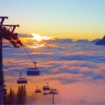 Über einem wolkenverhangenen Tal schwebende Skiliftsessel bei Sonnenaufgang, im Hintergrund sind die Berggipfel zu sehen.