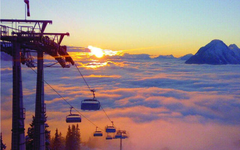 Über einem wolkenverhangenen Tal schwebende Skiliftsessel bei Sonnenaufgang, im Hintergrund sind die Berggipfel zu sehen.
