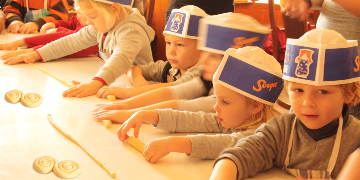 Mehrere kleine Kinder mit Papierhüten und Schürzen formen während einer Backaktion auf einem Tisch Teig zu Spiralen.