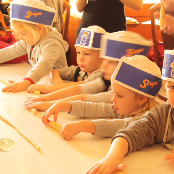 Mehrere kleine Kinder mit Papierhüten und Schürzen formen während einer Backaktion auf einem Tisch Teig zu Spiralen.