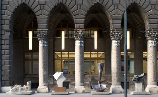 Eine Reihe von Steinsäulen steht vor einer bogenförmigen Gebäudefassade mit mehreren modernen Skulpturen und einem Schild mit der Aufschrift "MUSA" auf der linken Seite.