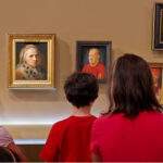 Drei Kinder und ein Erwachsener stehen vor einer Wand mit klassischen Porträtgemälden in einem Kunstmuseum. Der Erwachsene gestikuliert in Richtung der Kunstwerke, während die Kinder sie beobachten.