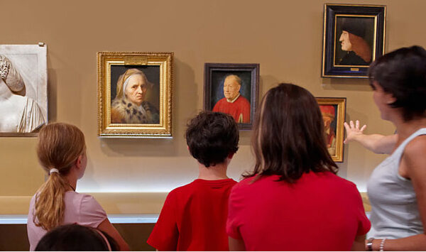 Drei Kinder und ein Erwachsener stehen vor einer Wand mit klassischen Porträtgemälden in einem Kunstmuseum. Der Erwachsene gestikuliert in Richtung der Kunstwerke, während die Kinder sie beobachten.