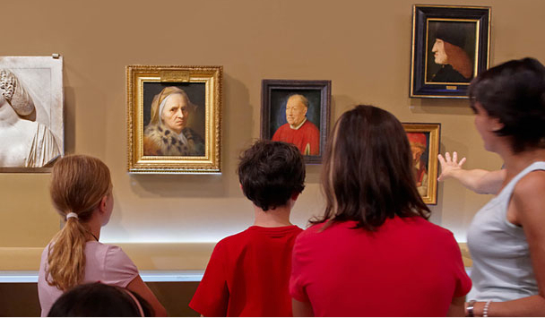 Drei Kinder und ein Erwachsener stehen vor einer Wand mit klassischen Porträtgemälden in einem Kunstmuseum. Der Erwachsene gestikuliert in Richtung der Kunstwerke, während die Kinder sie beobachten.