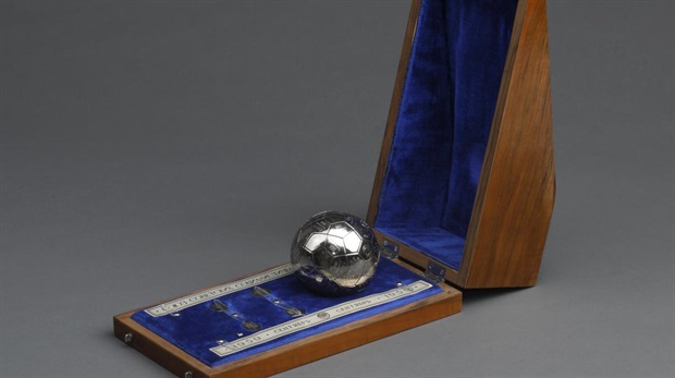 Eine silberne Fußballtrophäe in einer mit blauem Samt ausgekleideten Holzkiste, die offen auf einem grauen Hintergrund ausgestellt ist.